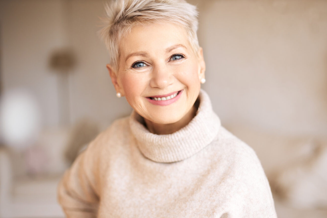 Stylish mature Caucasian female wearing cashmere turtle neck sweater looking at camera with joyful broad smile, showing perfect white teeth.